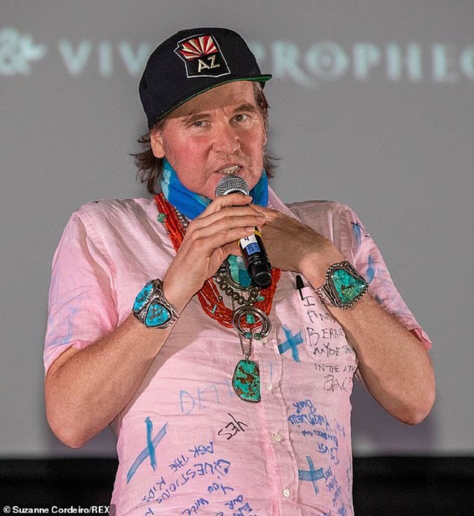 Val Kilmer speaks to a crowd while holding his hand to his neck, where he has a tracheostomy breathing aid