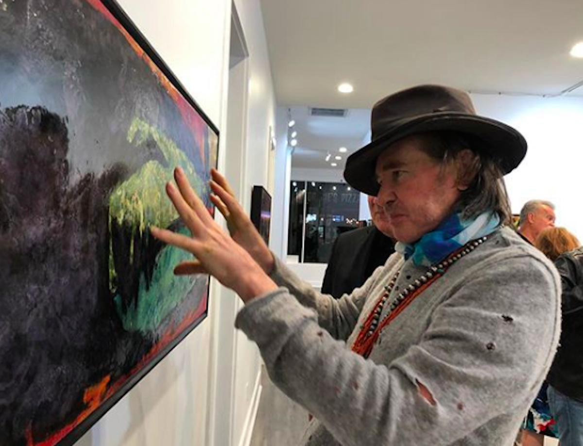 Val Kilmer with a hat posing in front of an art piece